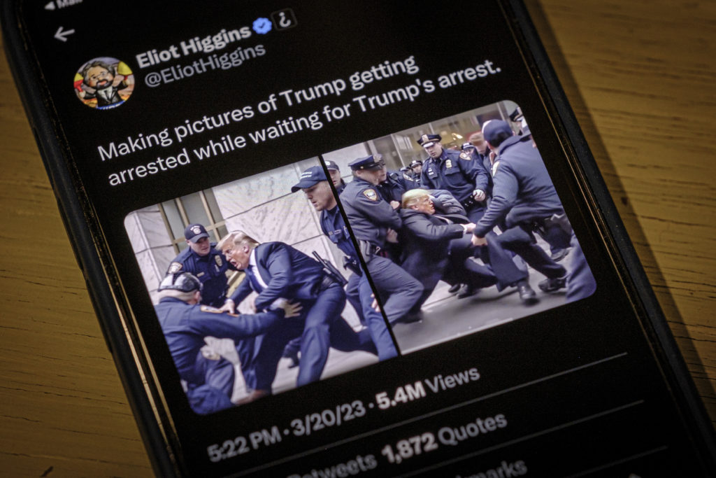 The image shows a smartphone displaying a tweet from a user named Eliot Higgins. The tweet seems to be commenting on the creation of fabricated images depicting a high-profile figure's arrest. The photo on the phone's screen shows several people in police uniforms holding down and arresting an individual resembling a public figure. The display suggests a discussion about digitally manipulated images or the anticipation of a significant event involving this public figure.